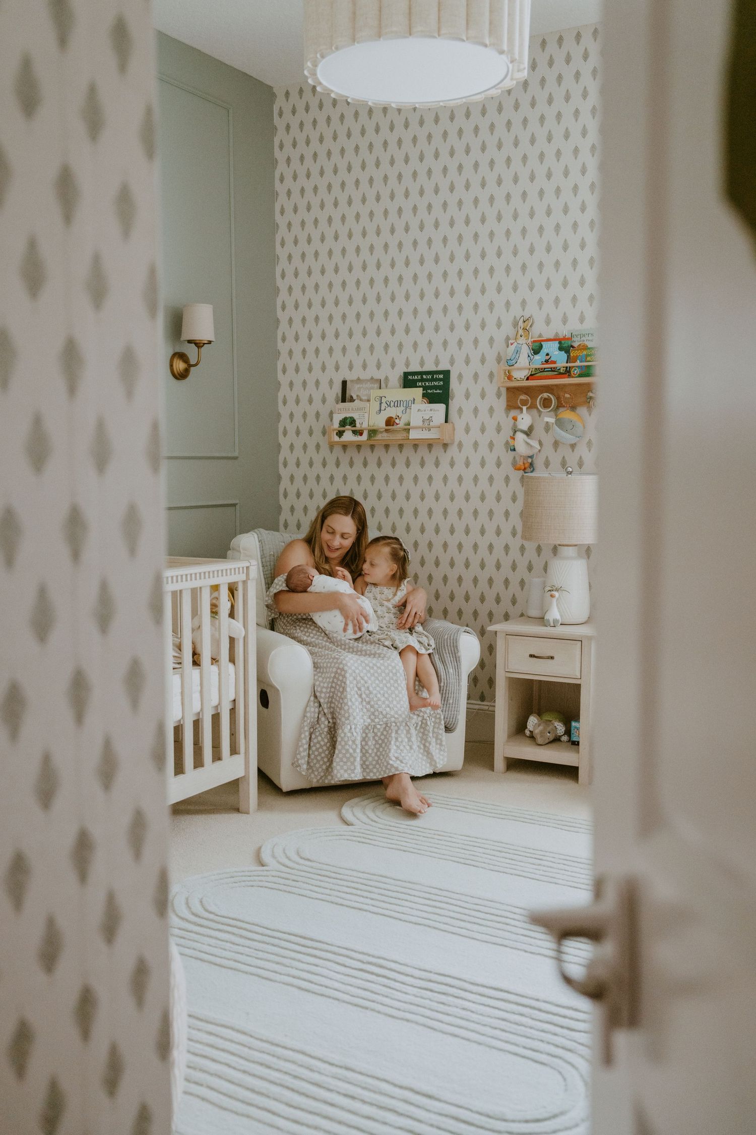 A cozy nursery room with striped area rug, patterned wallpaper, white crib, and large drum pendant light fixture.