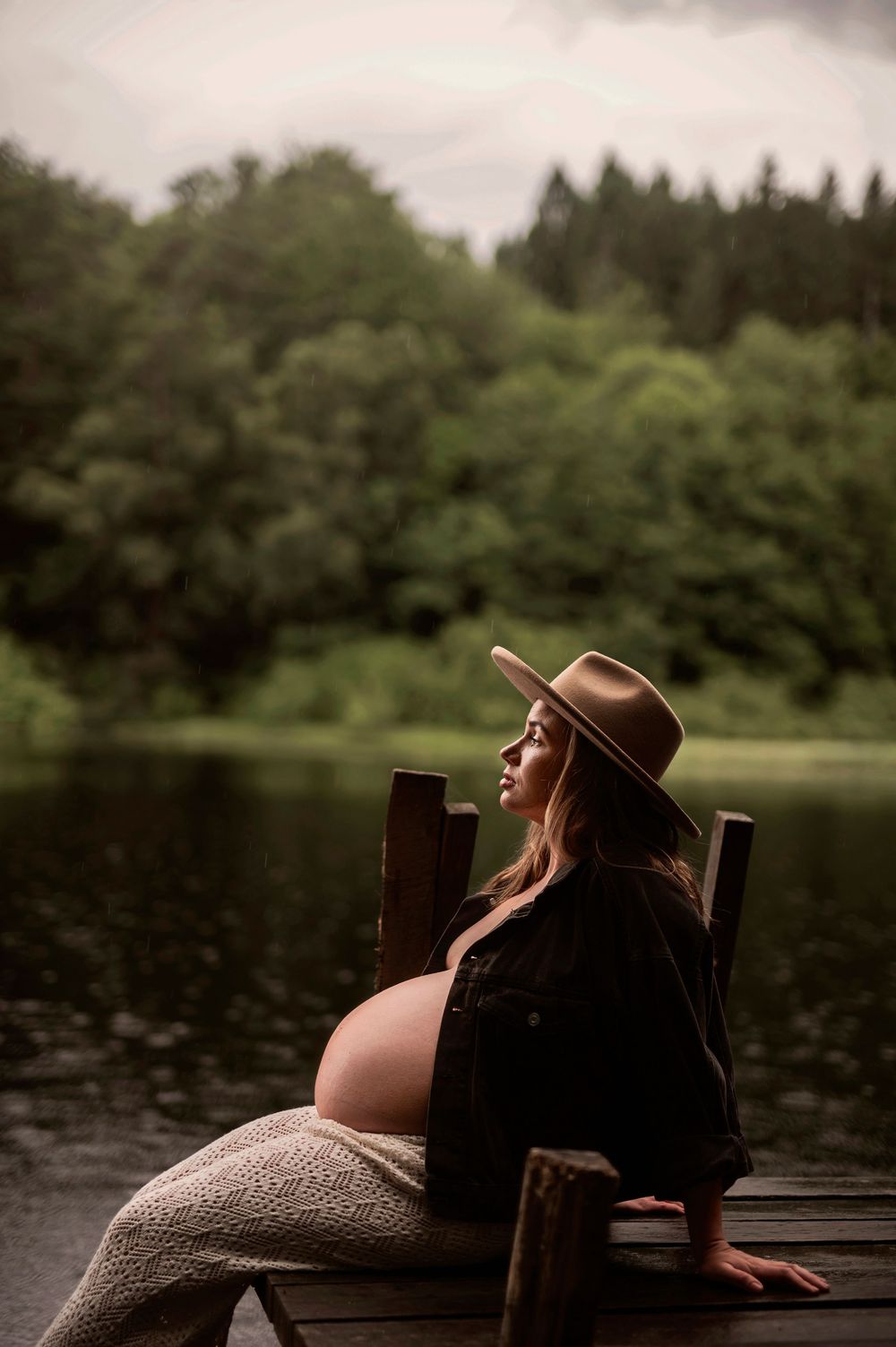 Gravid kvinna i brun hatt sitter lugnt på en brygga vid en stilla sjö, omgiven av grönskande skog.