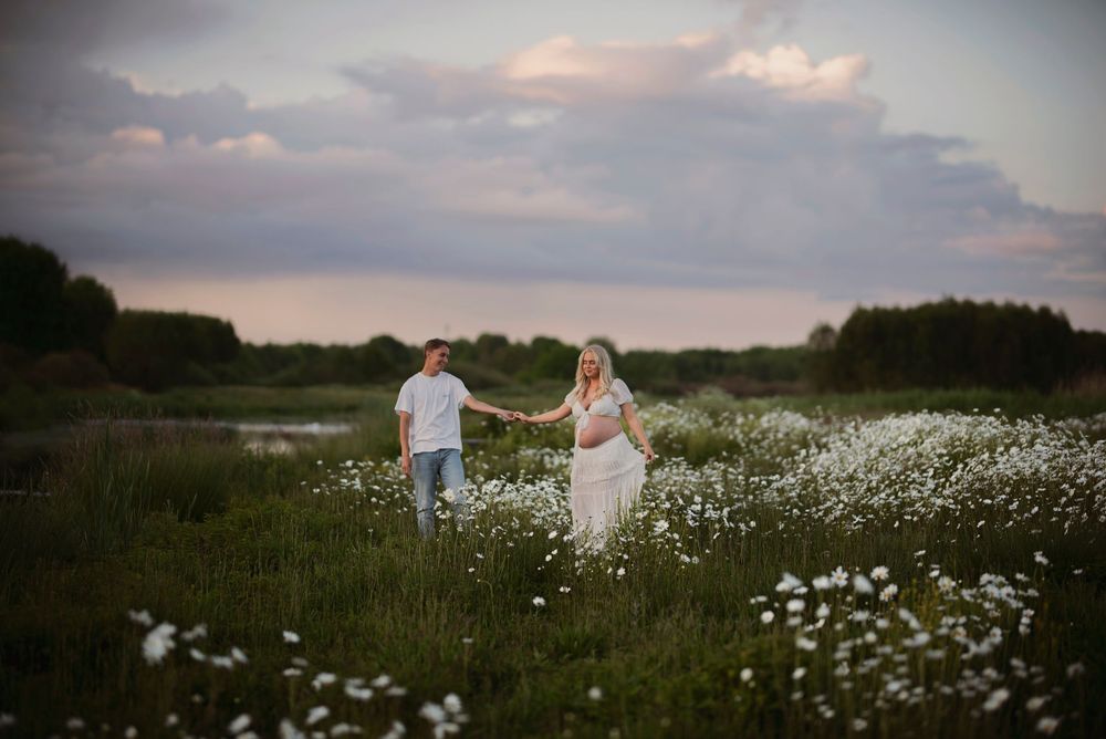 Blivande föräldrar som går hand i hand genom en blomsteräng i solnedgången, omgivna av  vita blommor.