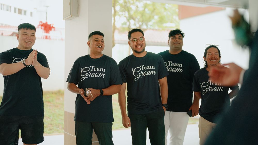A group of people wearing matching black Team Groom t-shirts laughing together at what appears to be a wedding event.