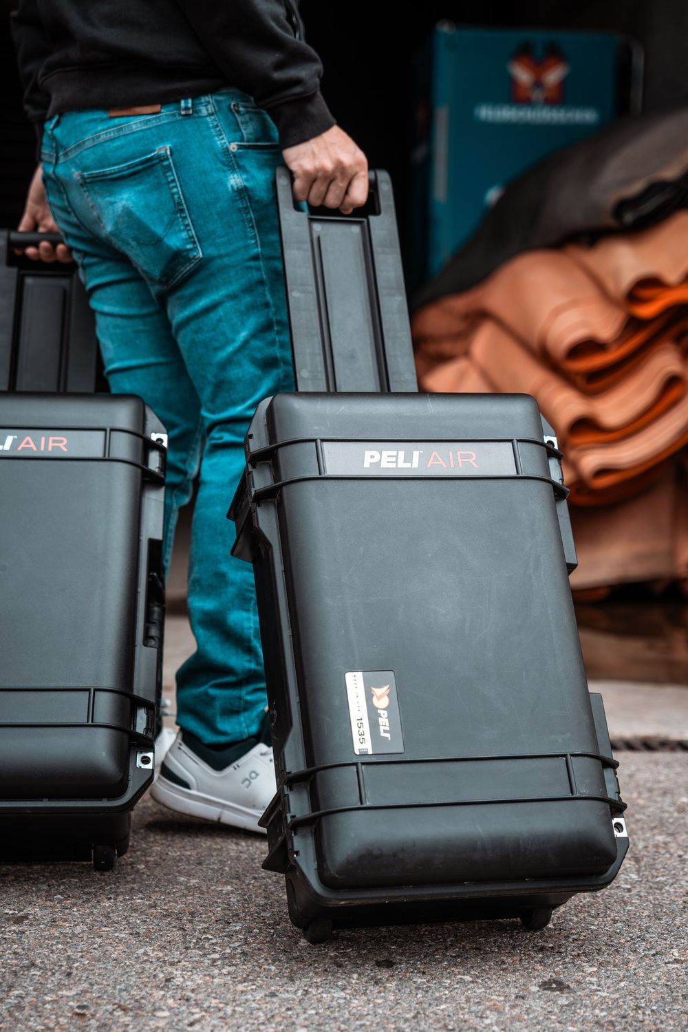 Grey and black Peli hard cases stacked on ground next to person wearing blue jeans.