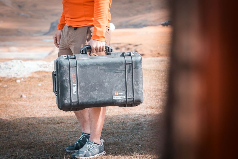 Person carrying a black camera equipment case outdoors on a dirt surface near grass and trees.