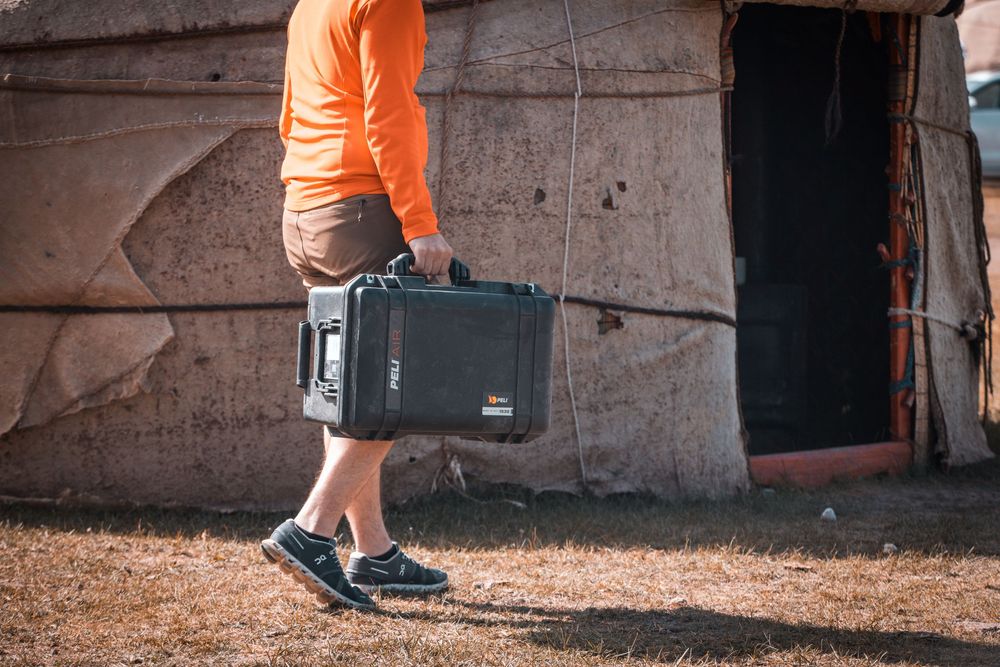 Orange-shirted person carrying a case while walking across dirt and concrete surfaces in multiple views.