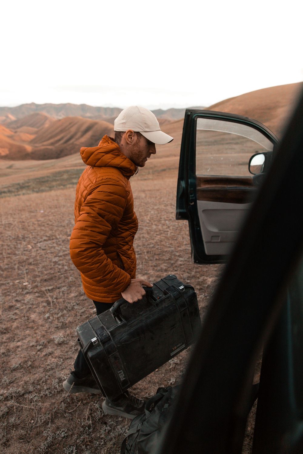 Person in orange jacket entering a car door in a desert landscape during sunset.