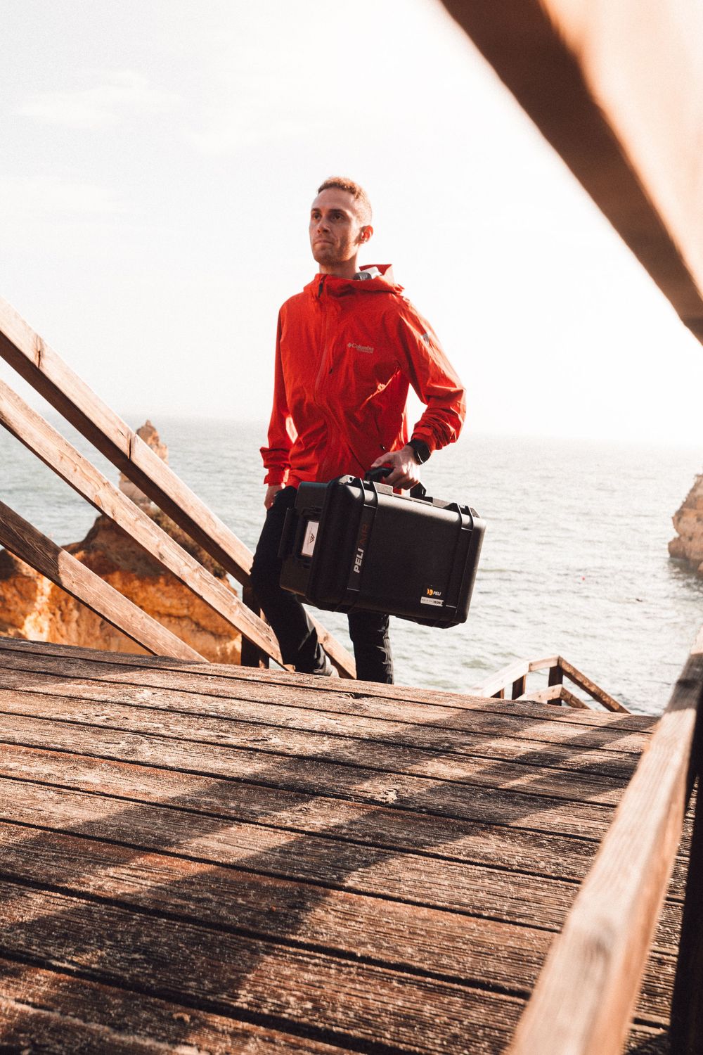 A person in red outerwear carrying a large case stands on wooden coastal boardwalk steps by the ocean.