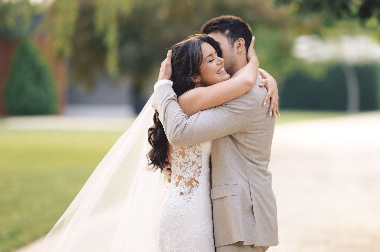 A bride and groom share a romantic moment by a lake, with the bride in white and groom in beige on a grassy shore.