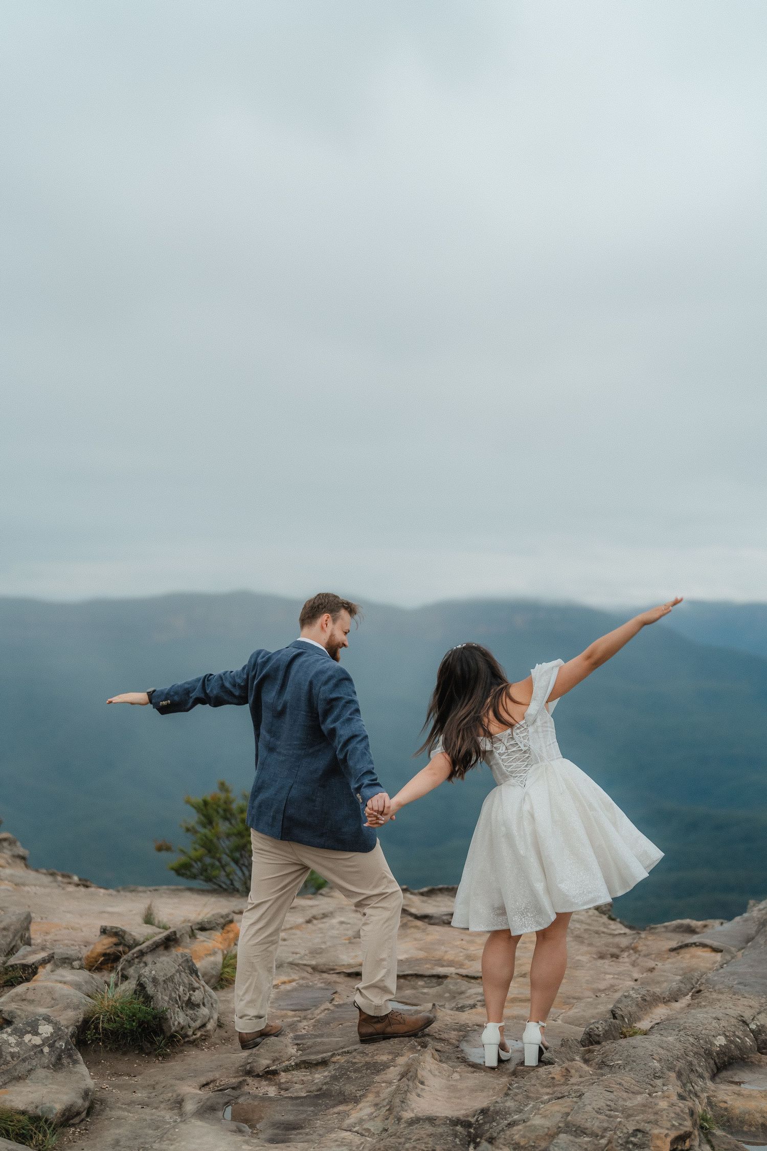 A playful couple strikes a balancing pose on rocky mountain terrain against a cloudy landscape.