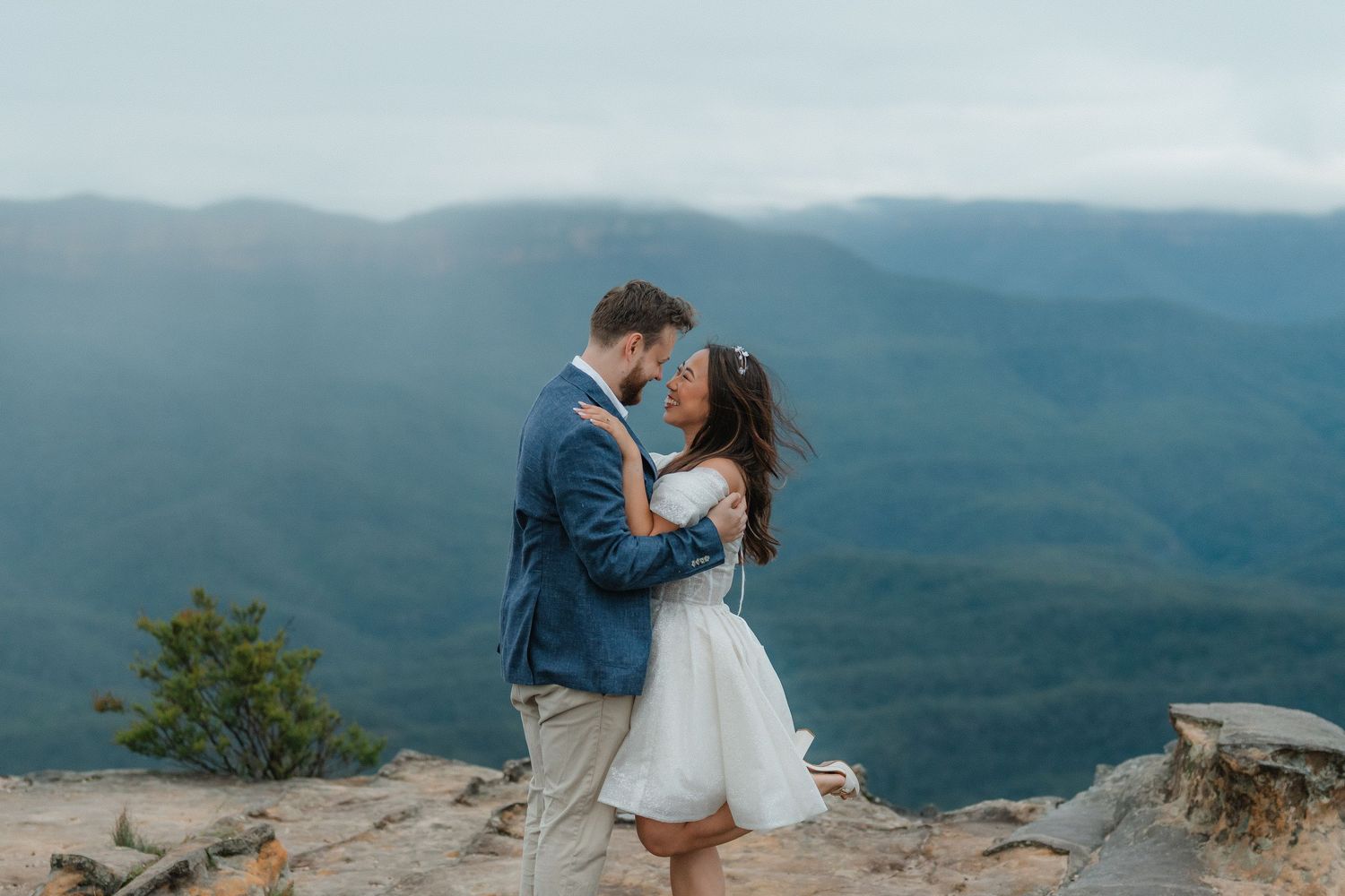 A couple embraces on a mountain overlook with sweeping forest views and misty peaks in the background.