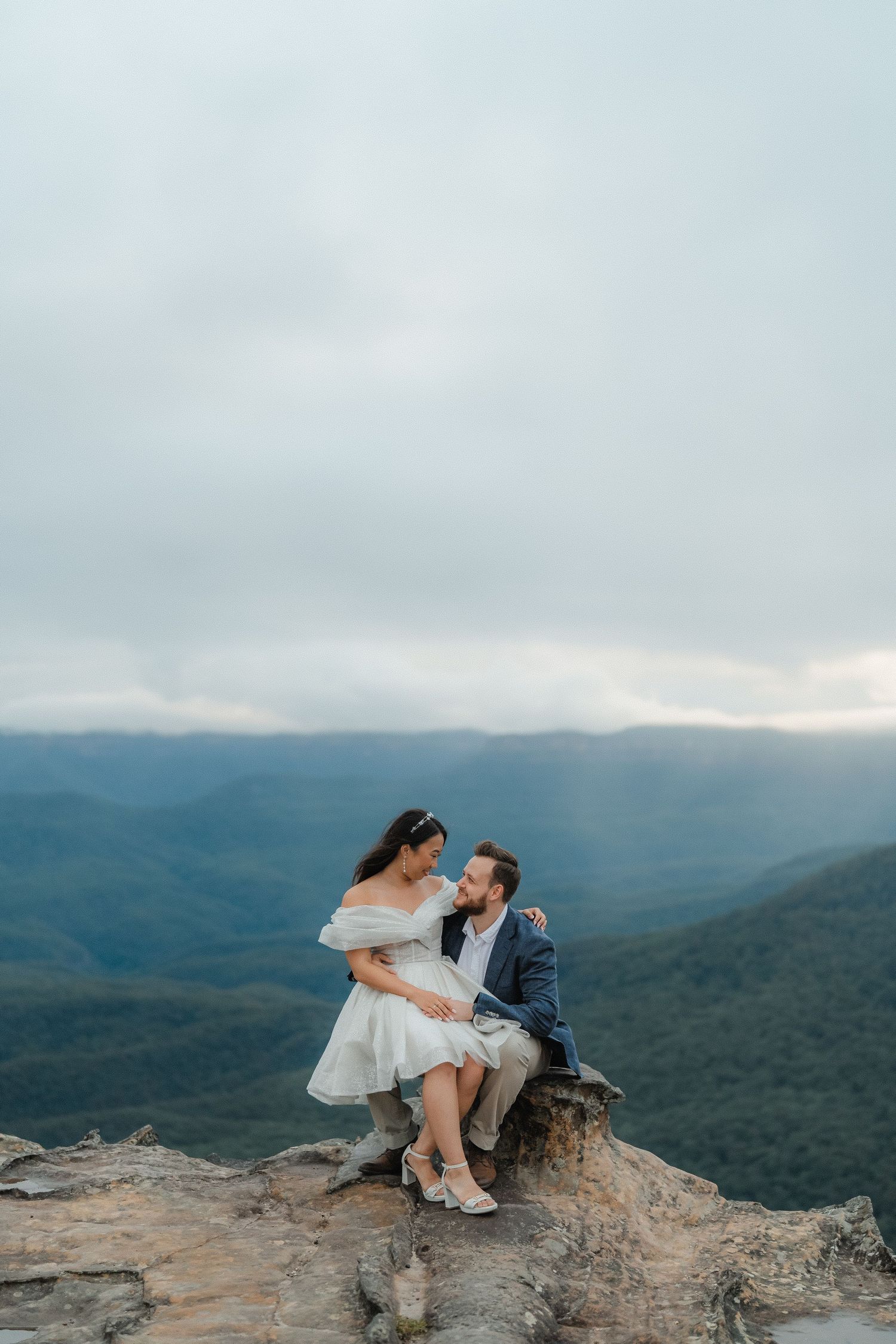 Couple in elegant attire sharing intimate moment on mountain cliff edge.