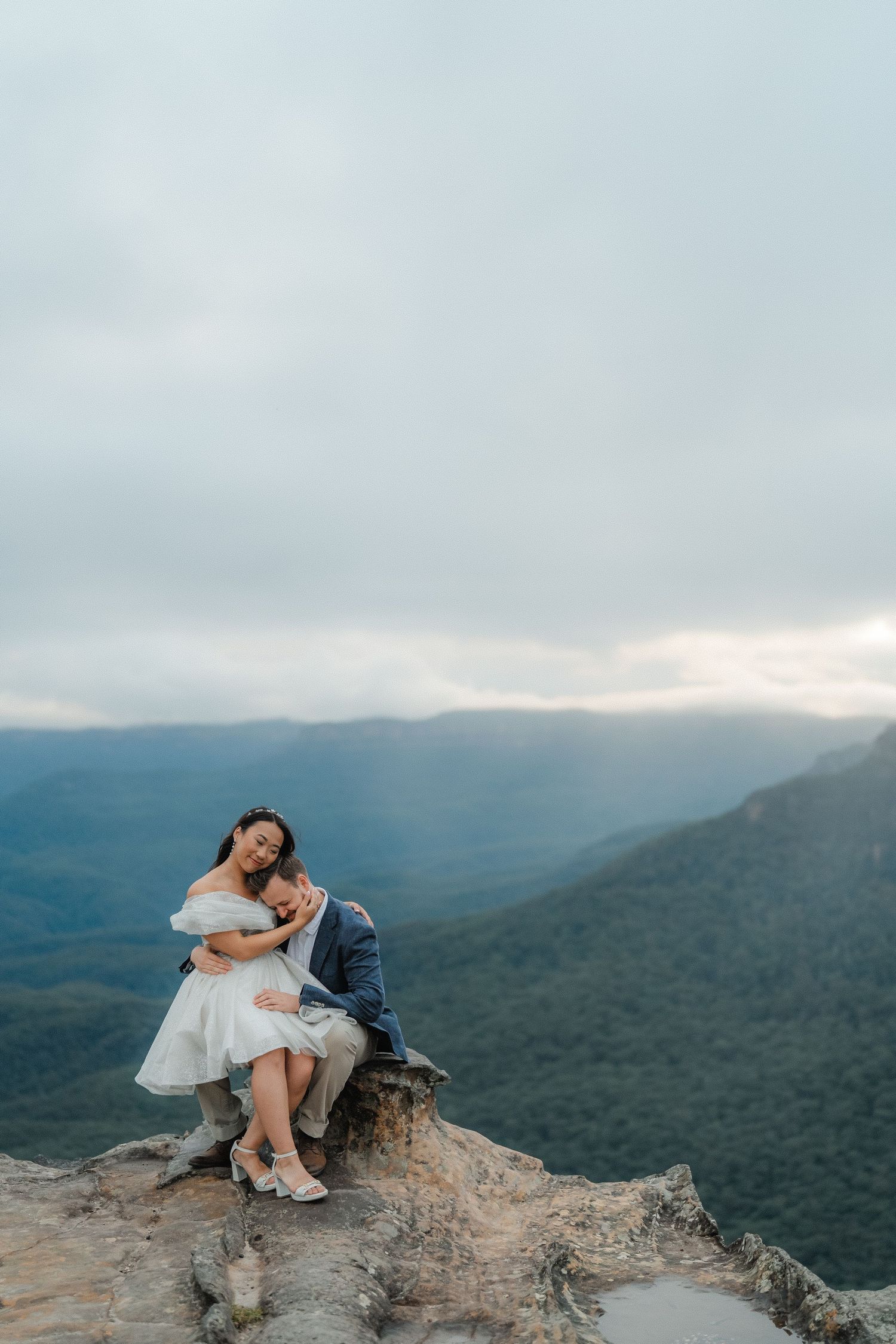 A couple embraces while sitting on a rocky cliff edge overlooking a misty mountain landscape.