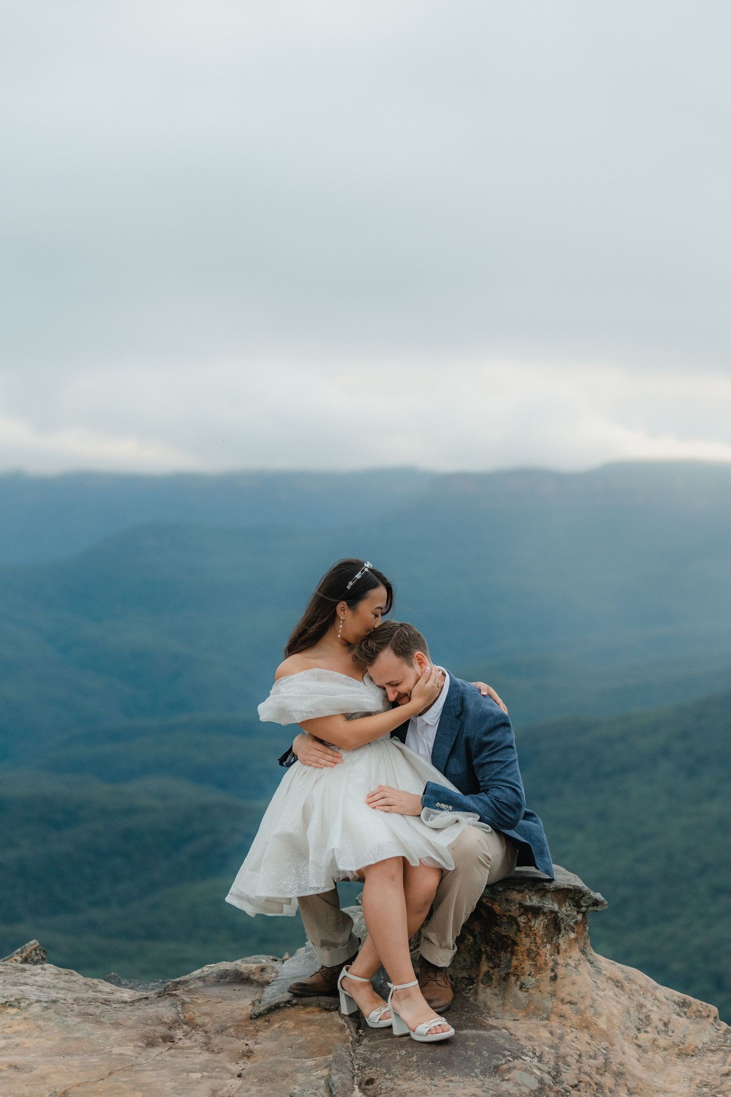 Couple sits together on rocky mountain ledge overlooking misty valley views.