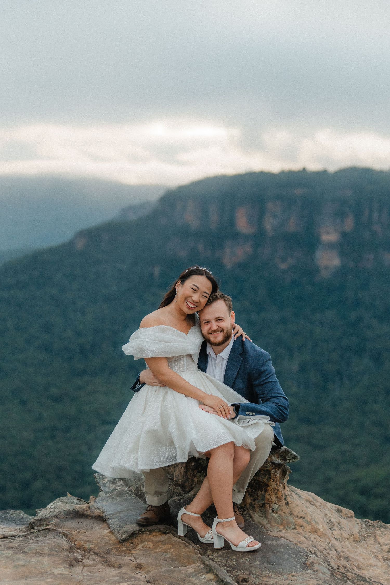 A happy couple shares a candid moment while sitting on a rocky outcrop with Blue Mountains in background.