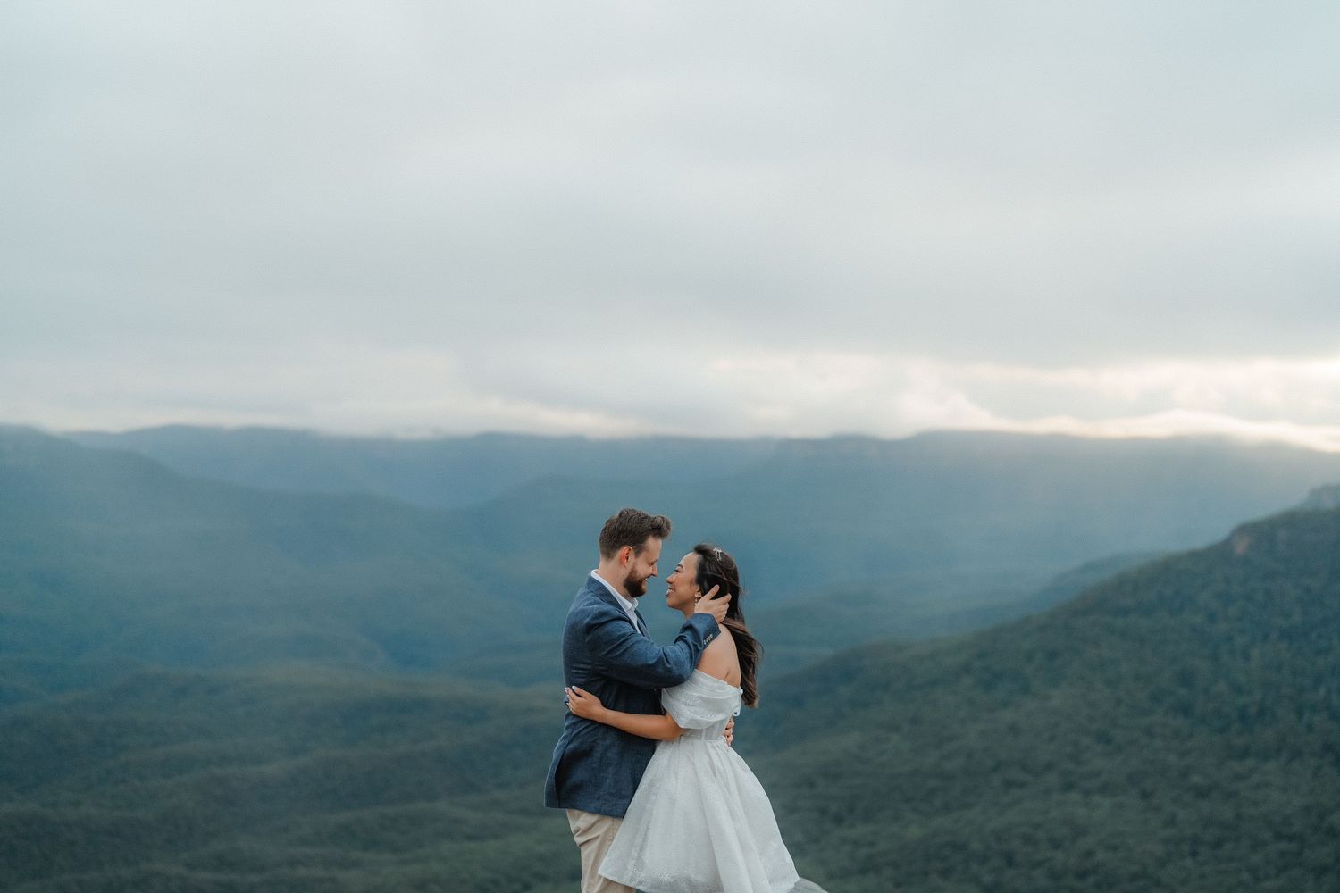 Couple in wedding attire standing on a mountaintop with a misty valley behind them.