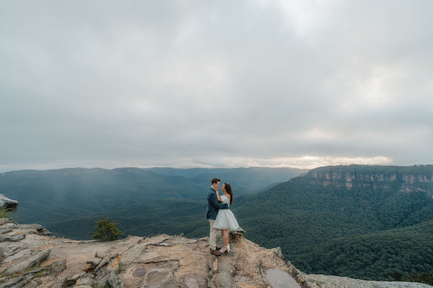 Couple shares an intimate moment while standing on a rocky mountain overlook at sunset.