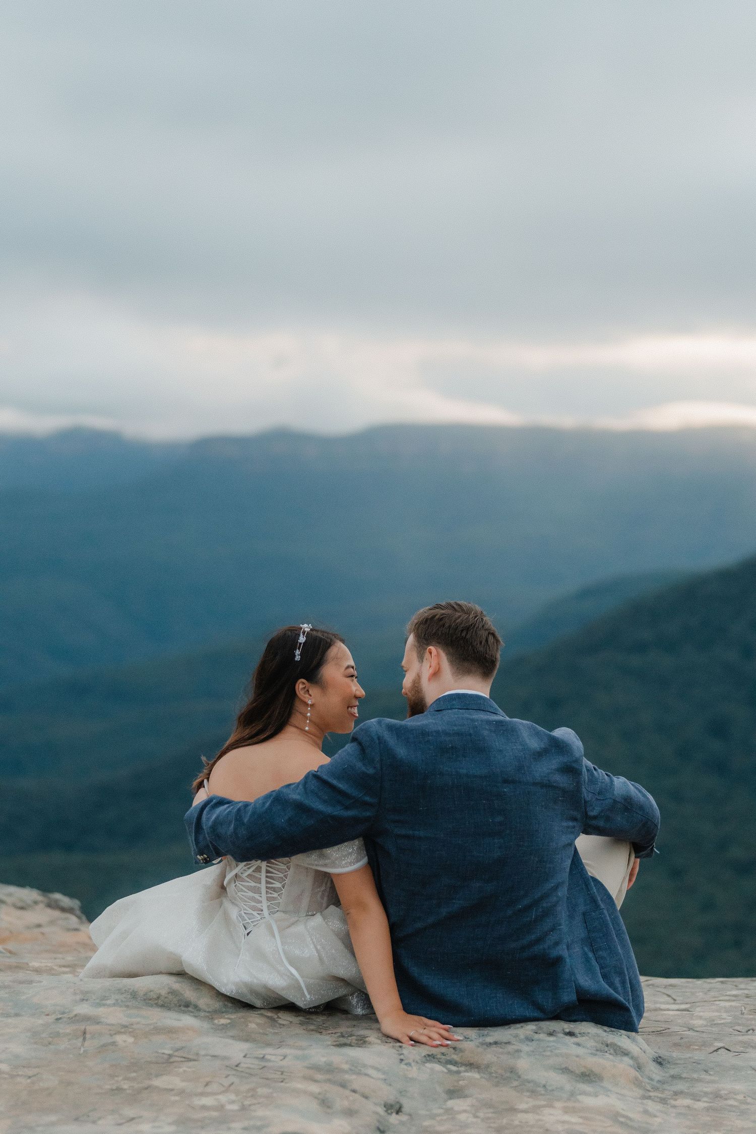 Two people sit together on a rocky mountain ledge enjoying the scenic valley views below.