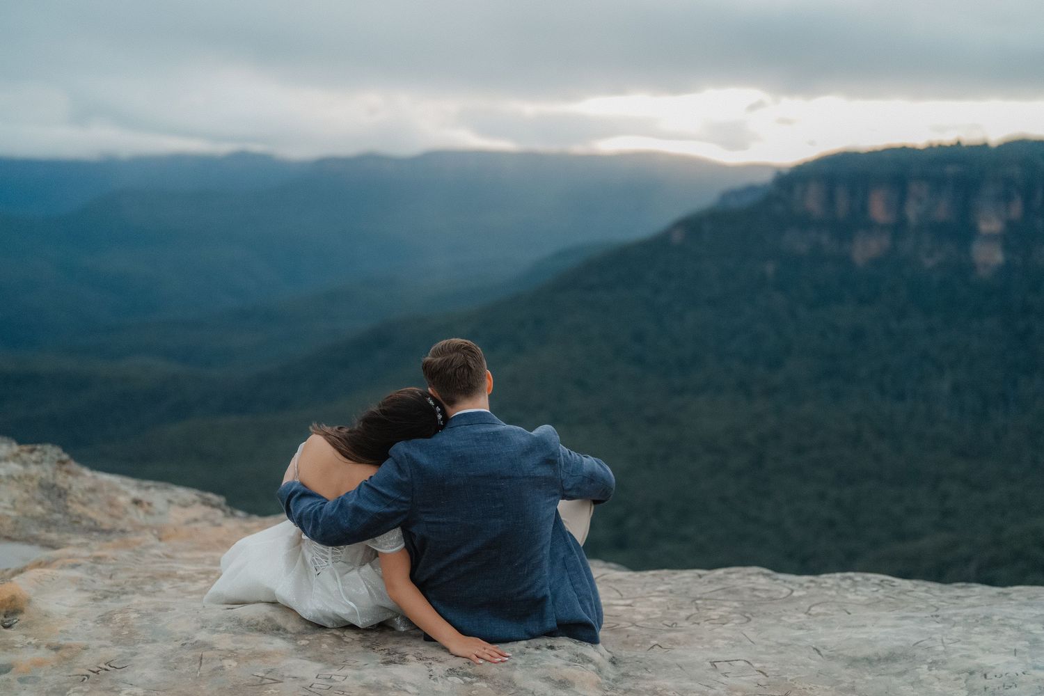 A couple sits close together on a mountain cliff edge overlooking a scenic forested valley.