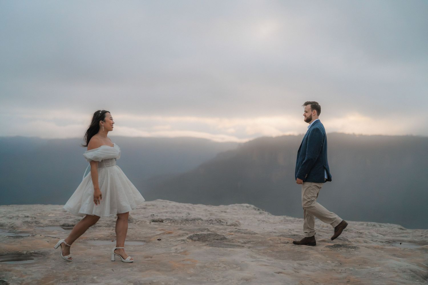 A couple playfully dances on a rocky mountain ledge against a misty mountain landscape.