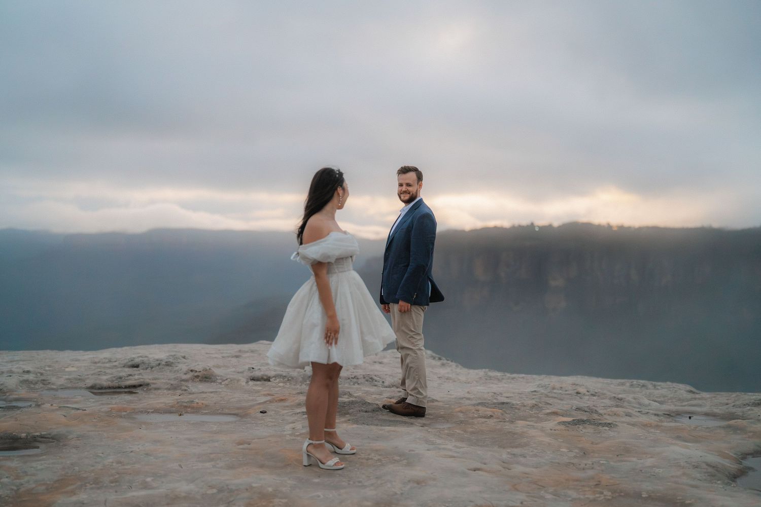 A couple dances playfully at sunset on a rocky mountain overlook with dramatic views of Blue Mountains in background.