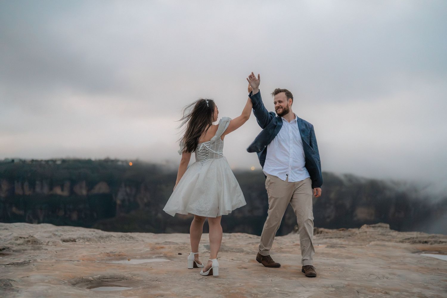 Playful dancing on mountain ledge with dramatic valley views and cloudy skies in background.