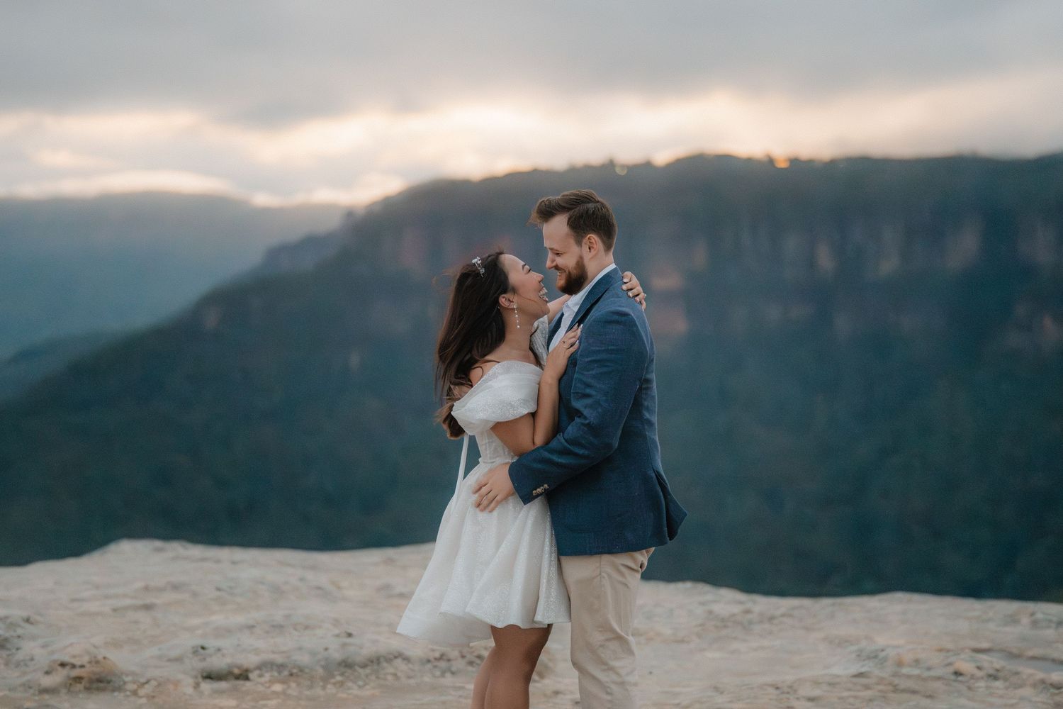 Couple embracing on a mountain cliff edge with dramatic valley views and cloudy skies.