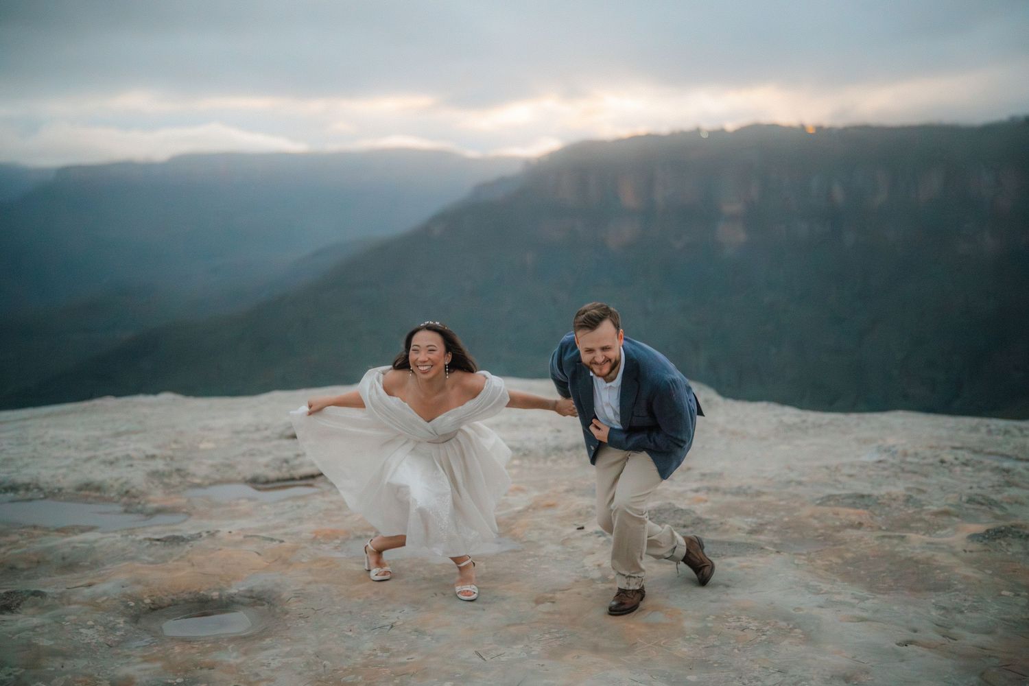 Couple poses playfully on rocky mountain ledge with sweeping valley views at dusk.