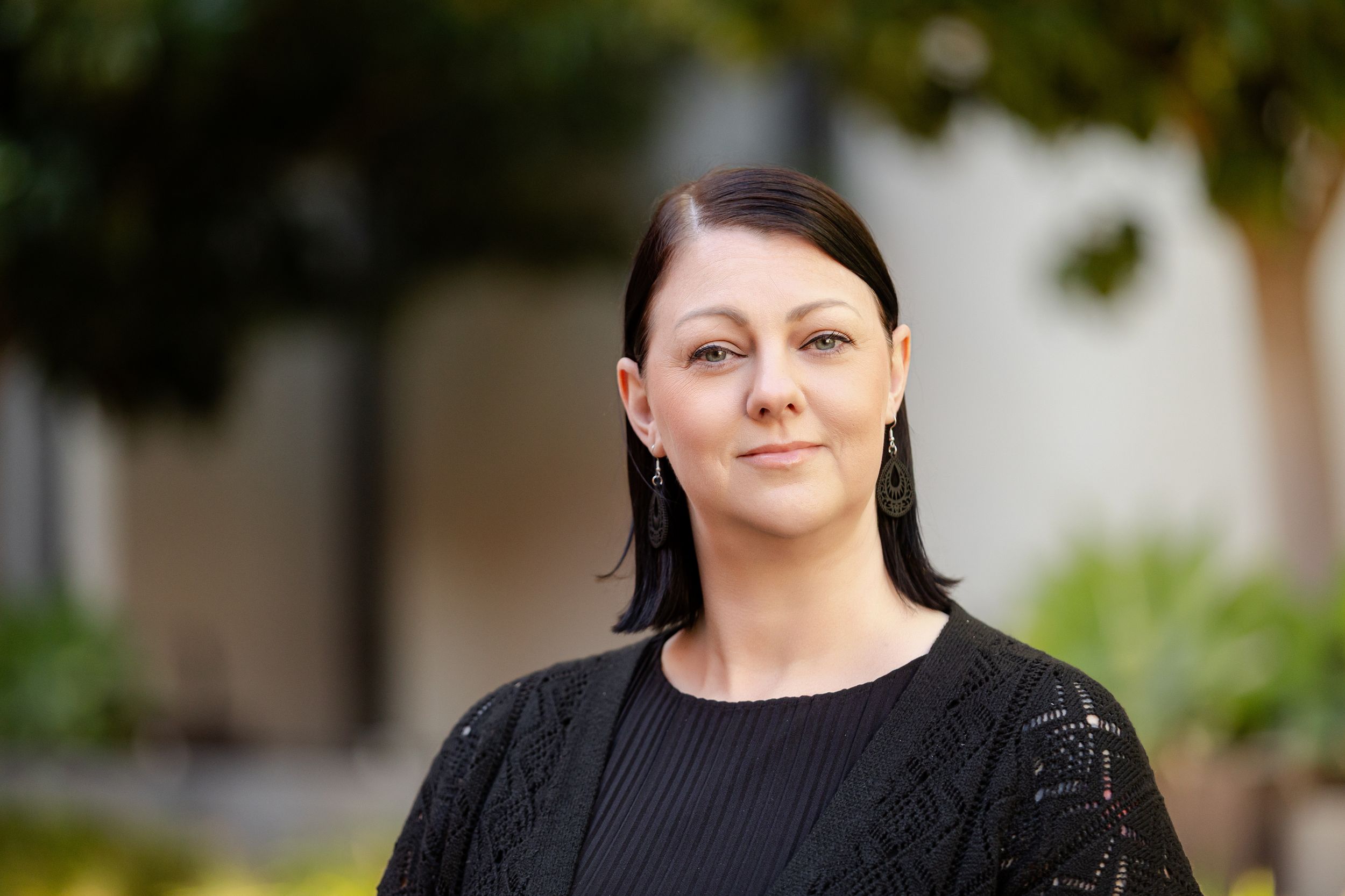 Professional headshot of someone in a black sweater outdoors with soft focus natural background.