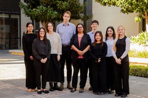 A professional team of colleagues in black and white business attire pose together outdoors in a sunlit courtyard.