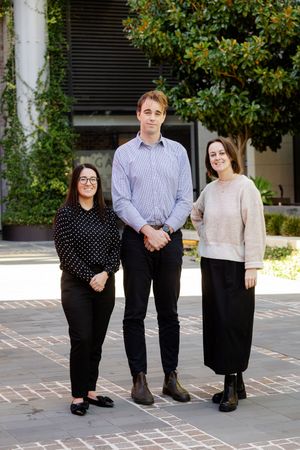 Three business professionals in casual office attire stand together on a brick courtyard.