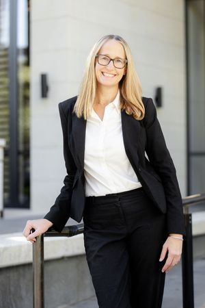 Professional in a black pantsuit and white blouse stands confidently against light building exterior.