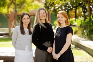 Three colleagues in professional attire pose outdoors in a sunlit garden setting.