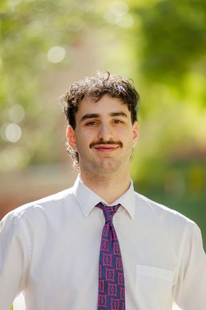 A professional headshot of someone wearing a white dress shirt and purple patterned tie against a blurred green background.