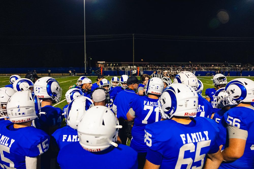 High school football team huddles under stadium lights wearing blue and white uniforms.