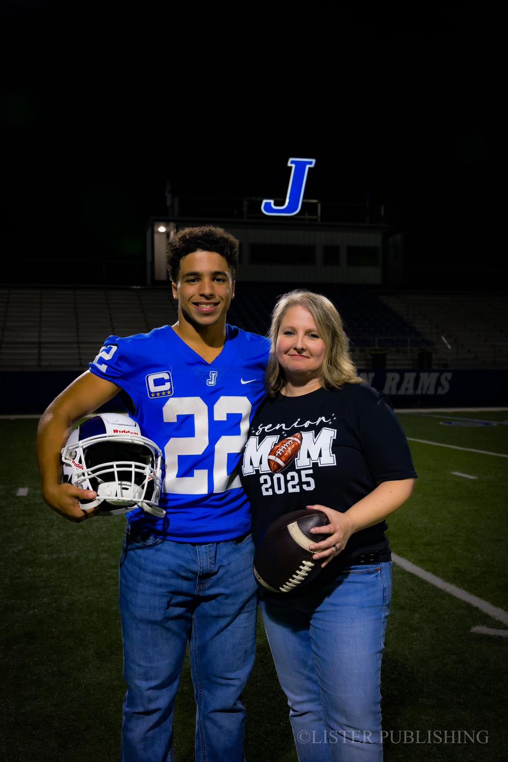 Student athlete and parent pose together on football field at night with team signage visible.
