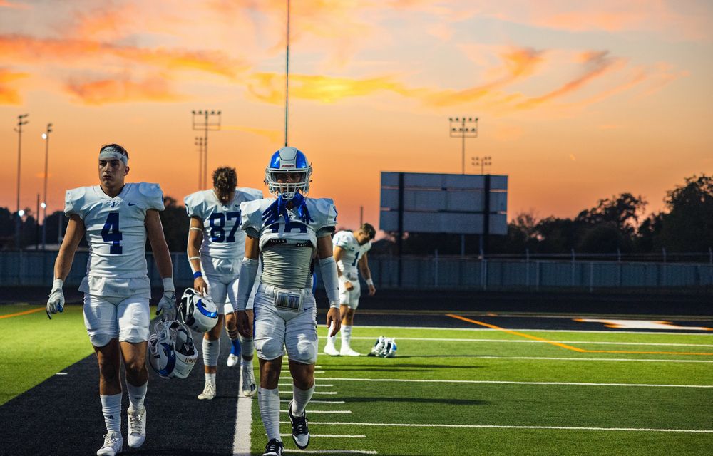 Football players walk across field during dramatic orange sunset with stadium lights illuminated.