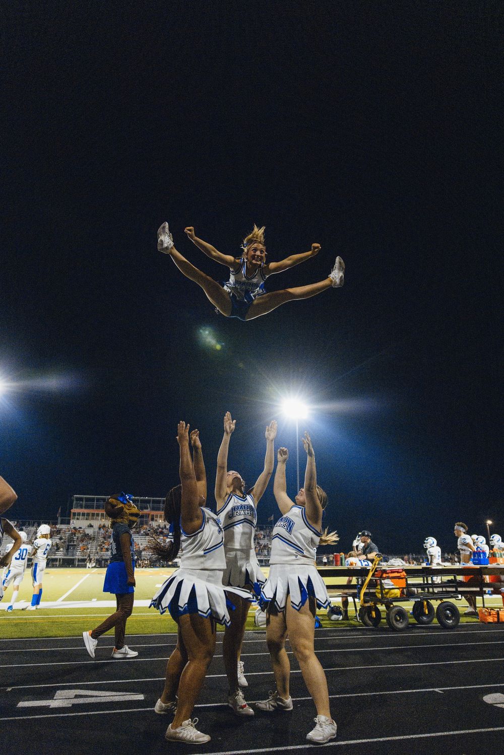 Cheerleaders perform aerial stunt during nighttime football game with stadium lights shining.