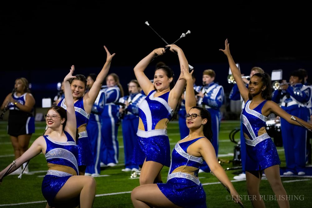 Cheerleaders in blue uniforms perform synchronized dance routine on football field at night.