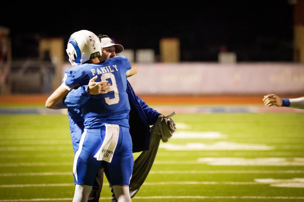 Football players embrace on field during game in blue uniforms.