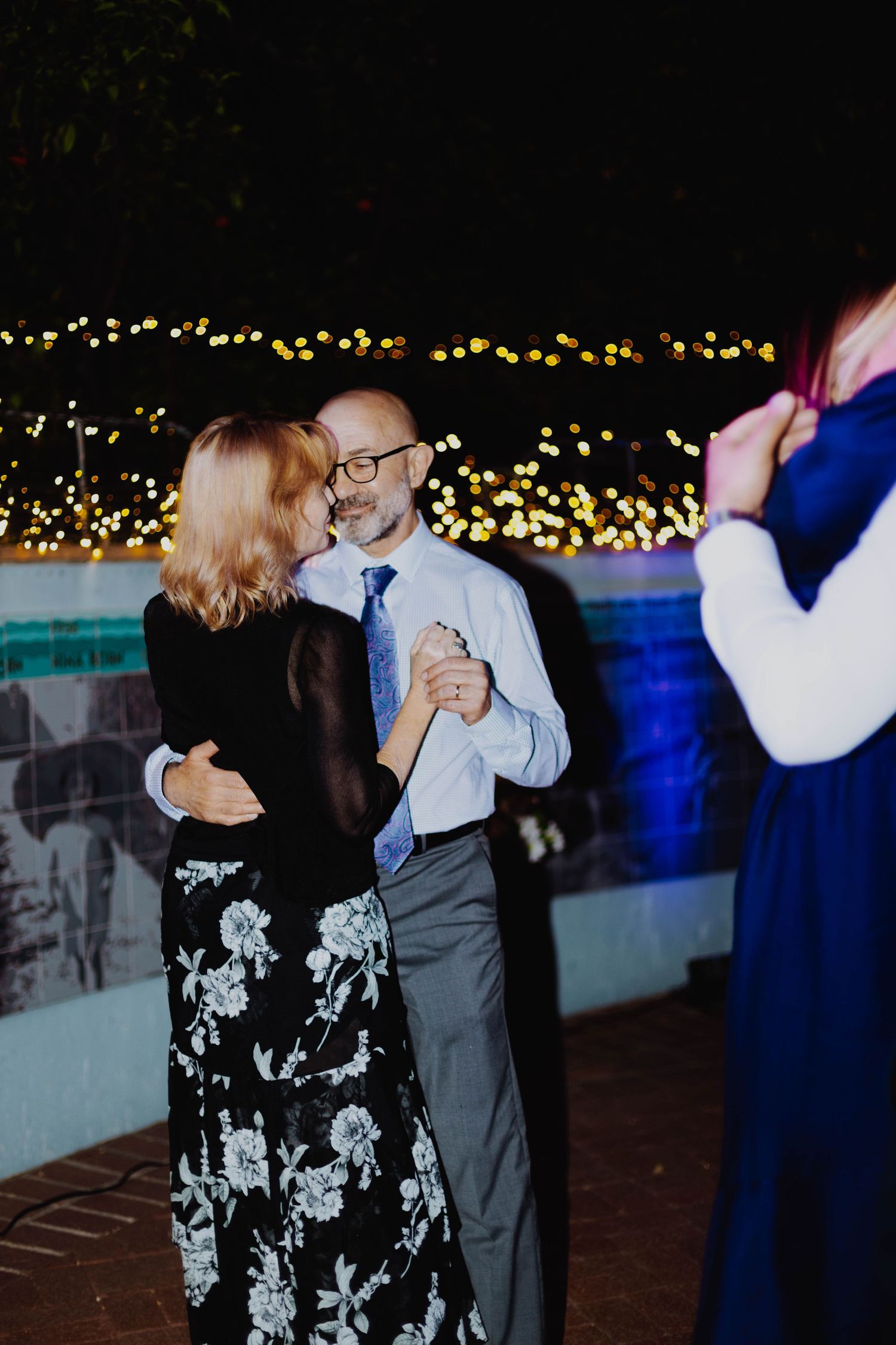 A couple dancing at an evening event with twinkling lights in the background and a floral dress.