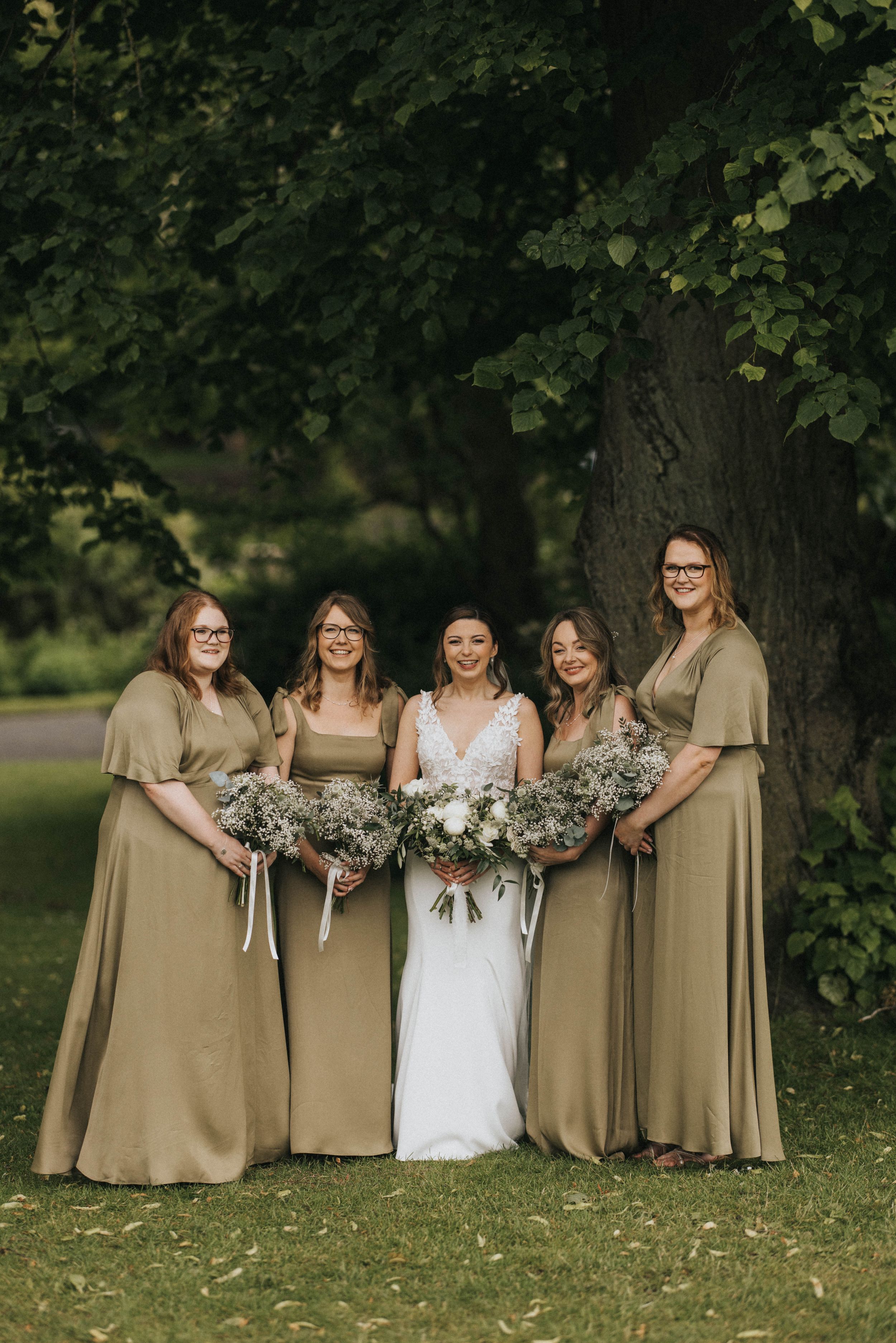 A group wearing tan bridesmaid dresses stands in a line embracing under green trees at an outdoor wedding ceremony.