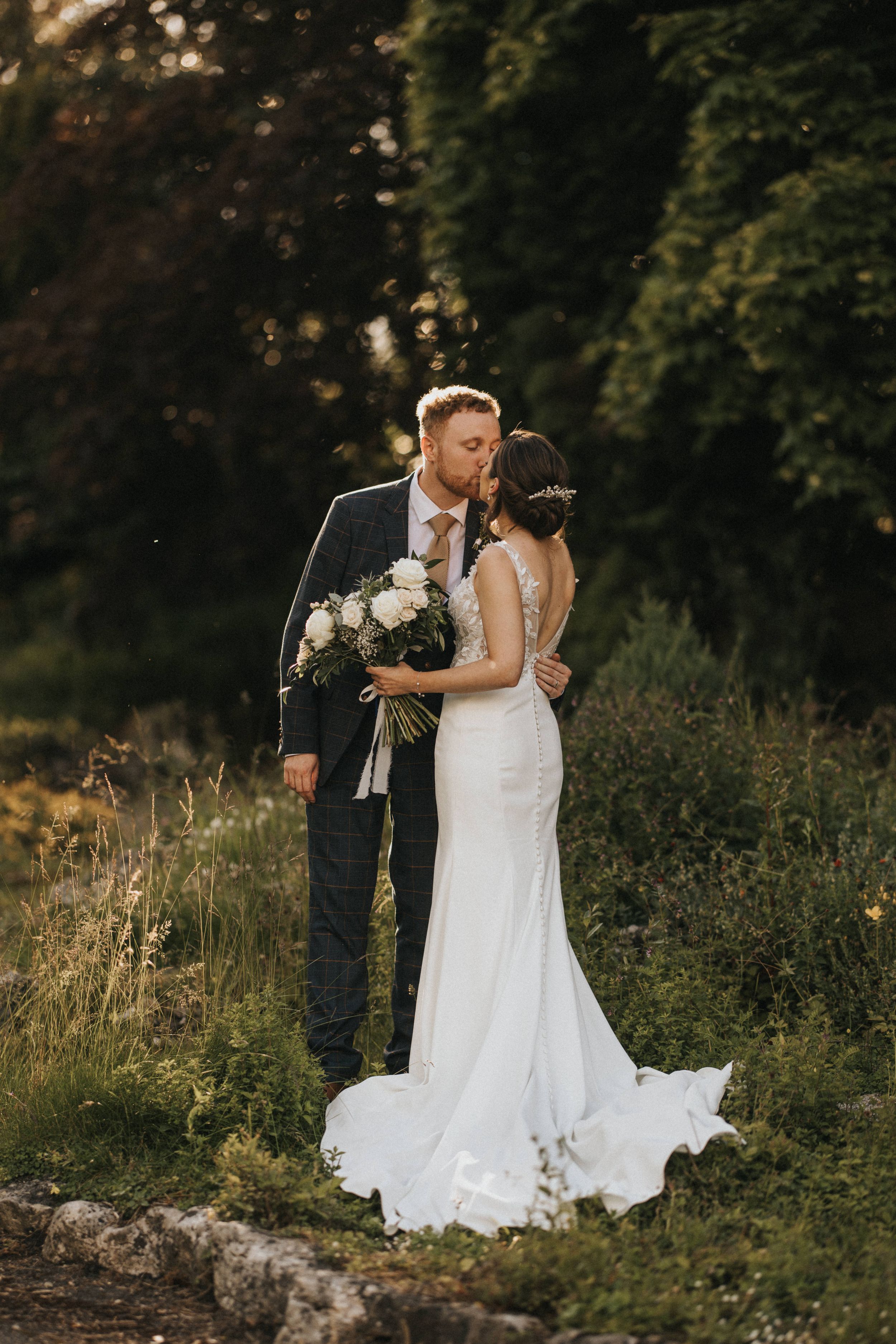 A newlywed couple embraces outdoors in natural lighting while holding a bouquet amidst greenery and wildflowers.