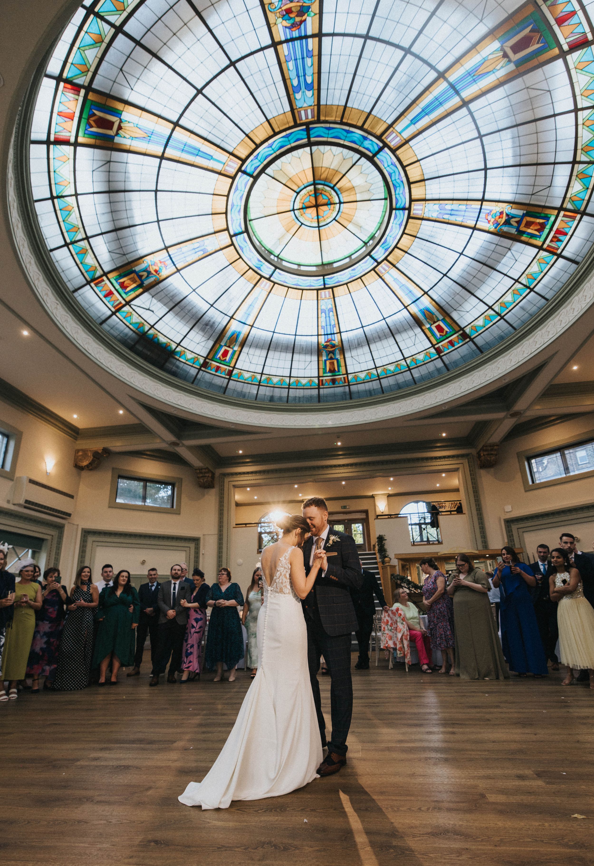 A wedding couple stands beneath an ornate stained glass dome ceiling in an elegant ballroom with guests surrounding them.