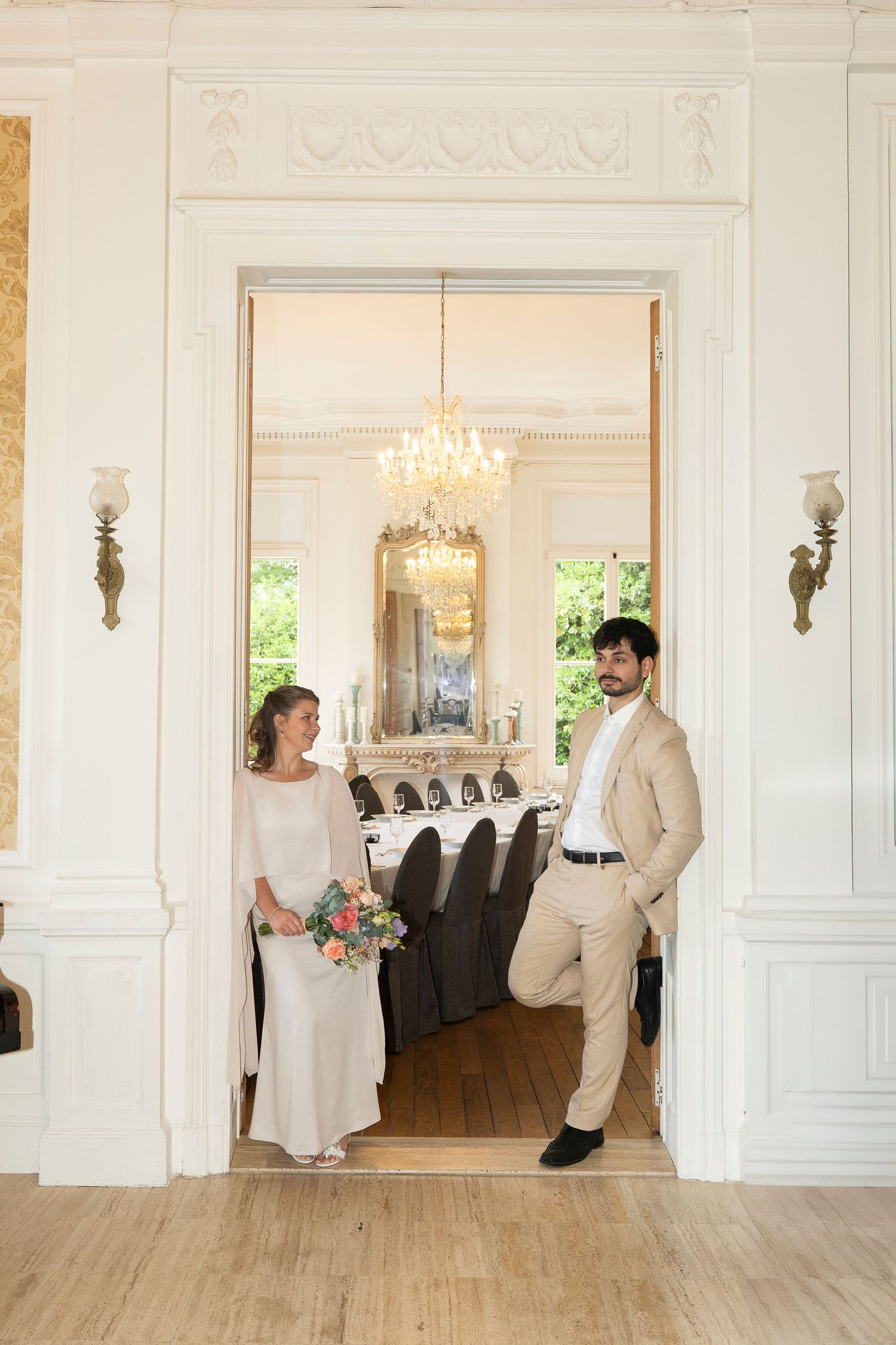 Romantic first look sequence between newlyweds in an elegant white ballroom with chandelier and hardwood floors.