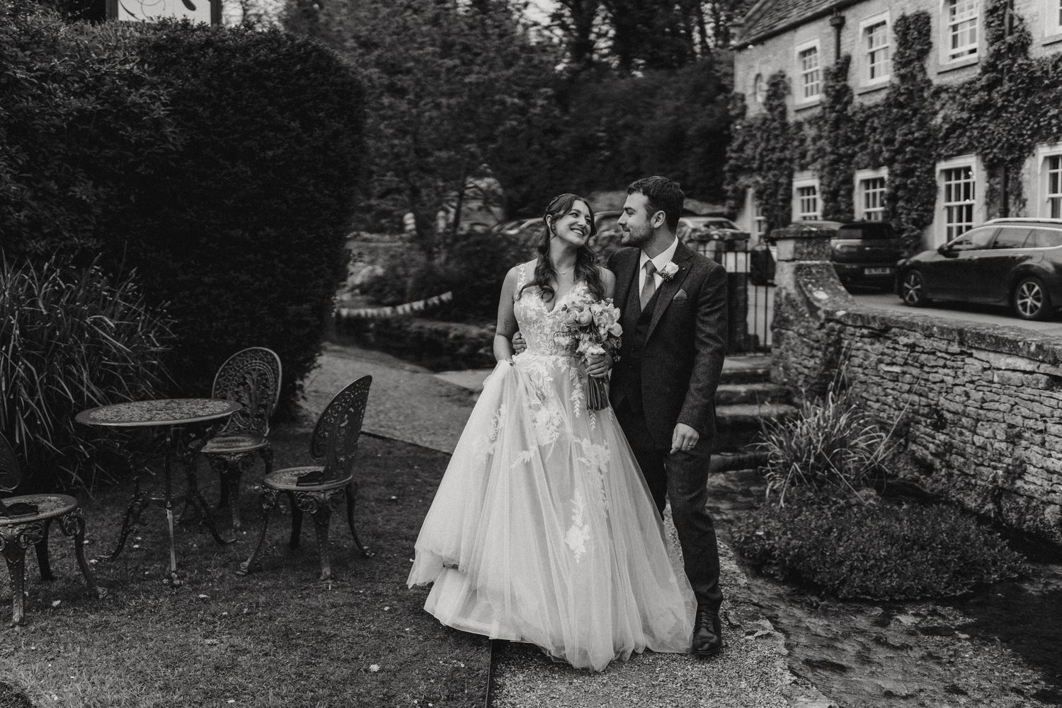 Elegant black and white wedding photo of a couple in formalwear standing in a garden outside a stone building.