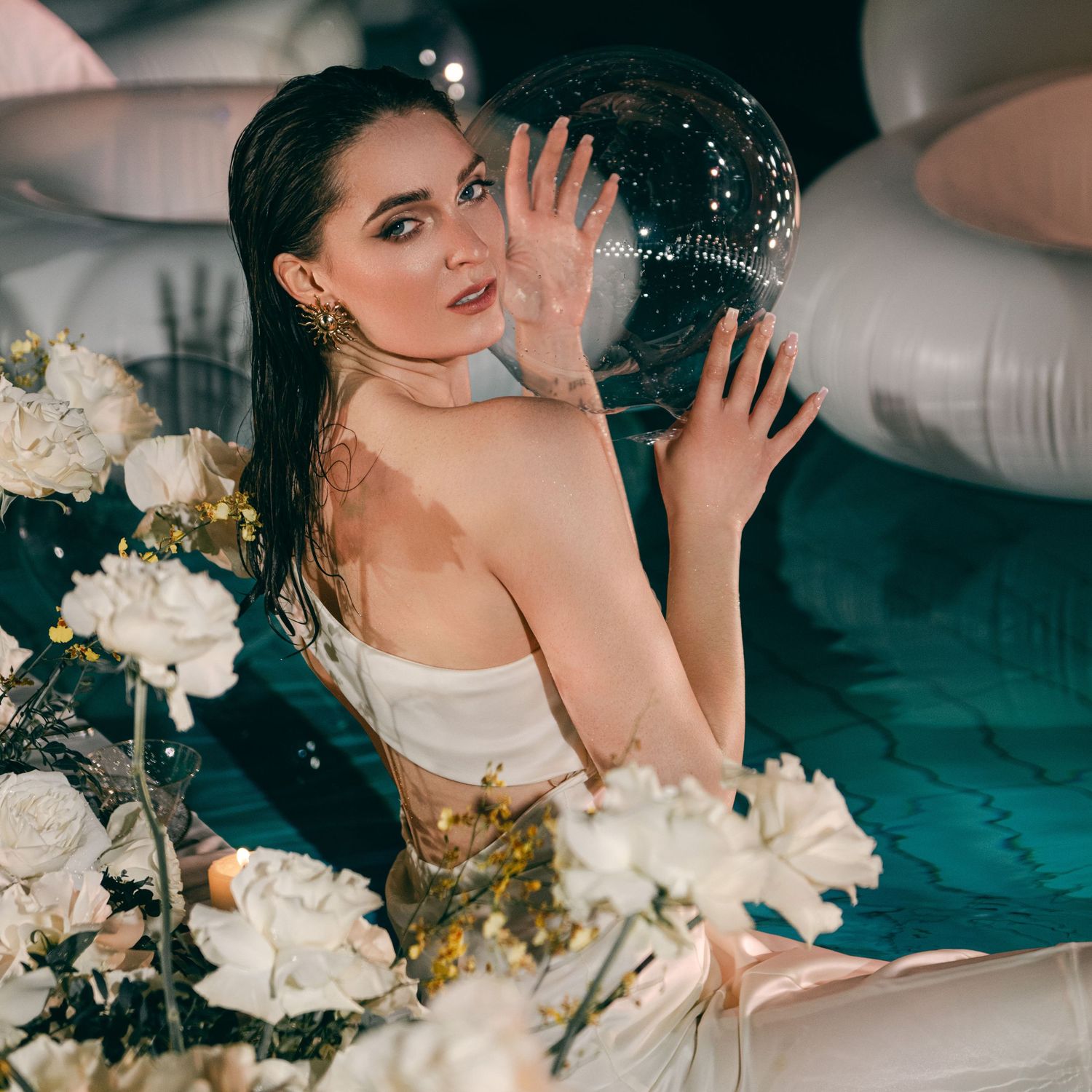 Woman in white dress poses with disco ball and white roses amid turquoise water and silver balloons.