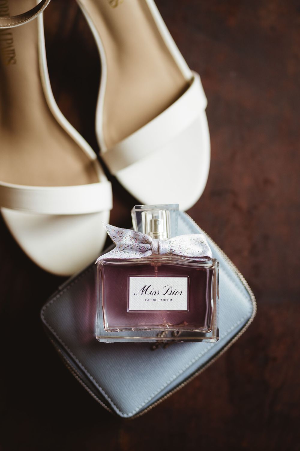 Elegant white bridal sandals and Miss Dior perfume bottle arranged on dark surface for wedding day detail shot.