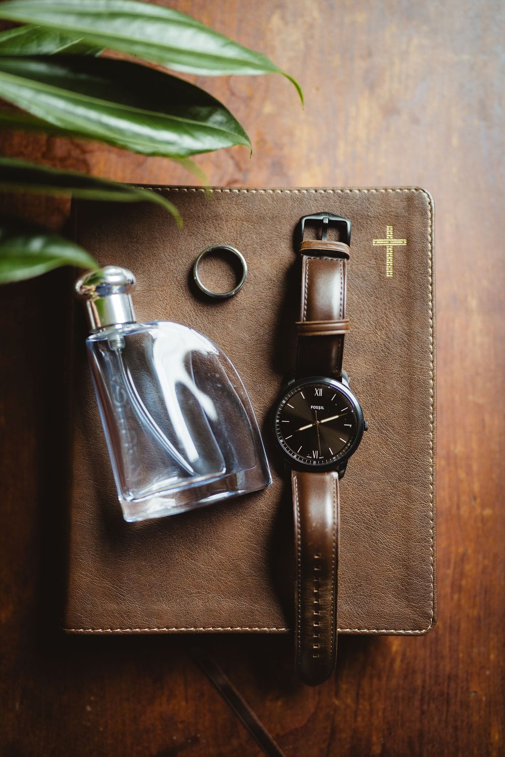 Stylish brown leather wallet with watch and glass perfume bottle arranged on wooden surface with subtle plant elements.