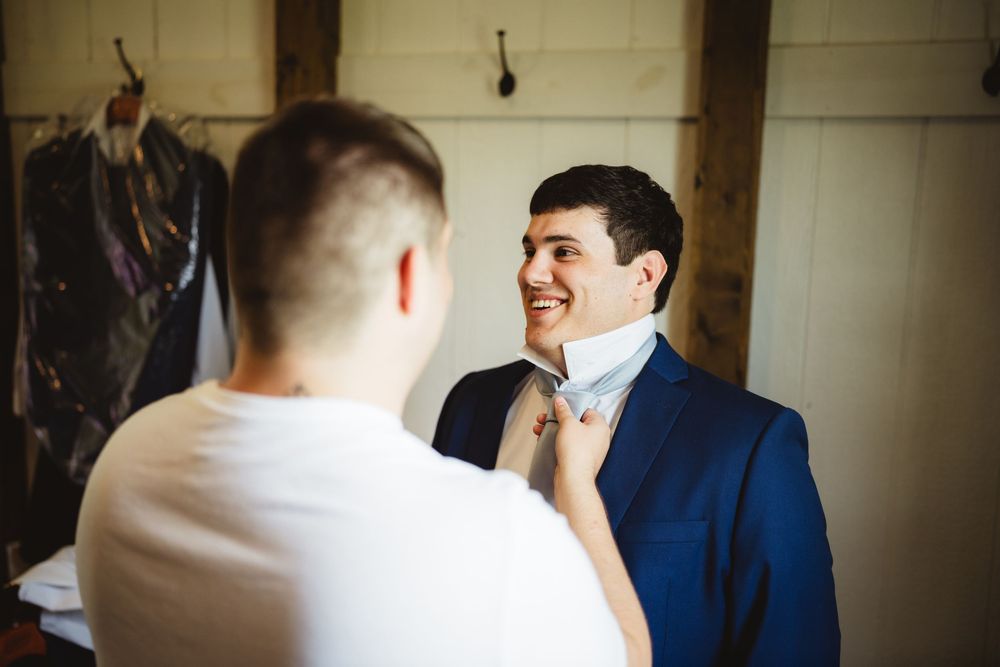 A groom in a navy blue suit smiles while someone adjusts his bow tie during wedding preparations.