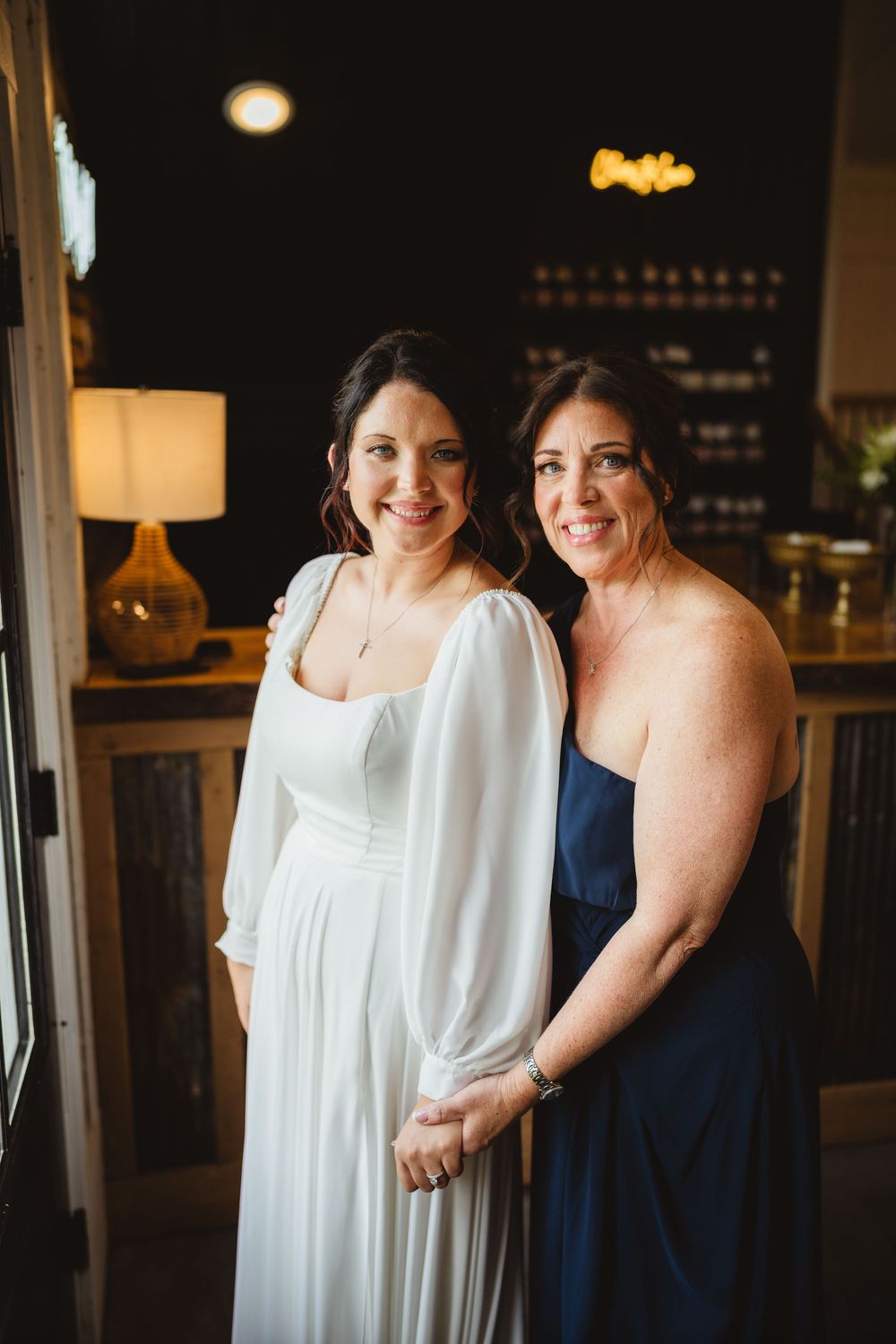 Two women wearing white and navy dresses pose together in dark room with warm lighting.