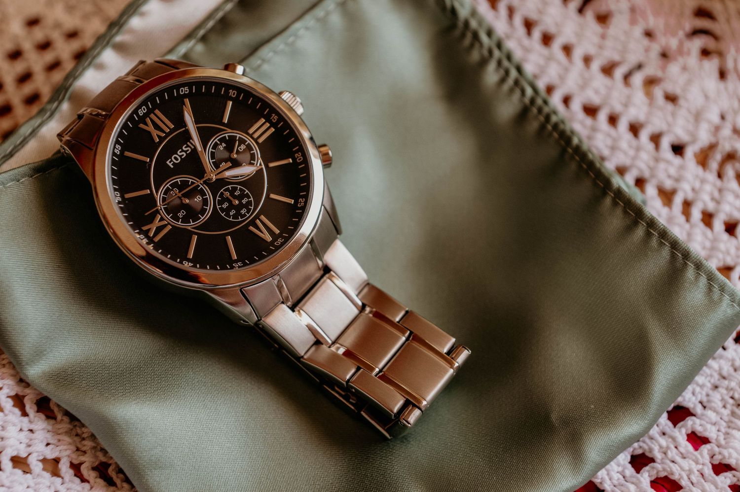Elegant rose gold chronograph watch with black dial and metal bracelet rests on sage green fabric with lace detail.