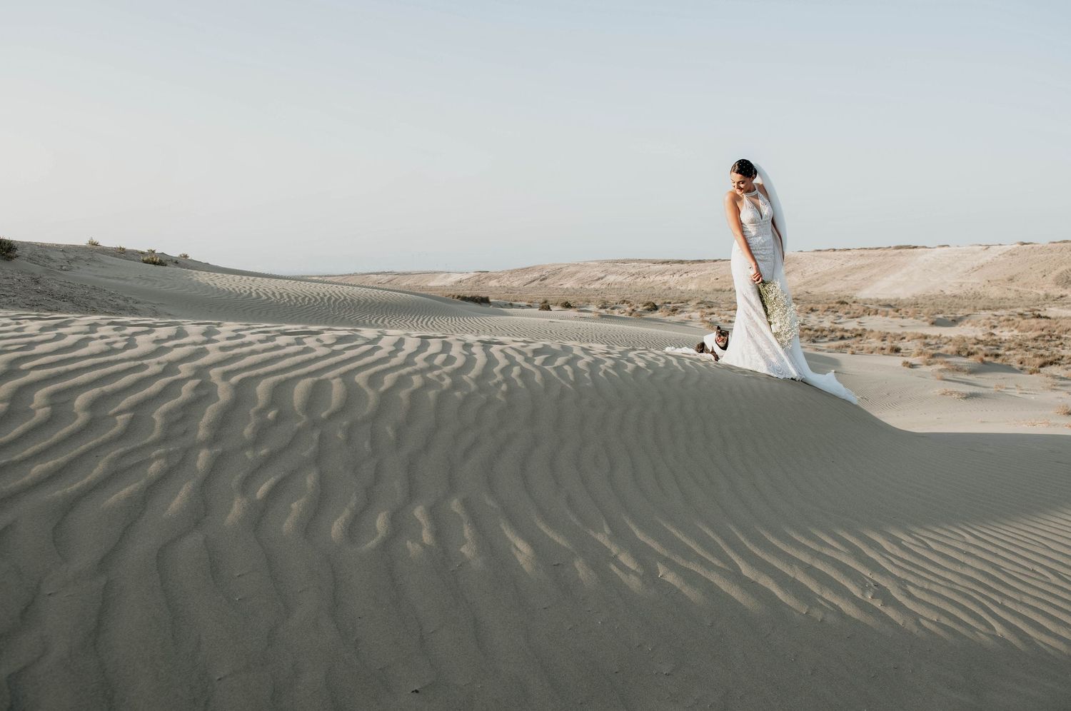 A wedding dress creates an elegant silhouette against rippling sand dunes at sunset.